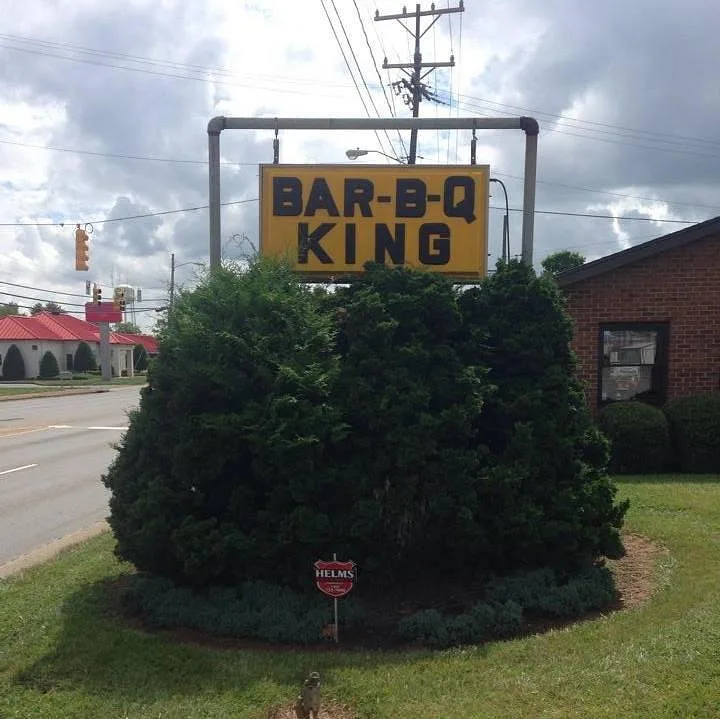 Classic yellow BAR-B-Q KING road sign on East Main St, Lincolnton, NC, framed by evergreen shrubs