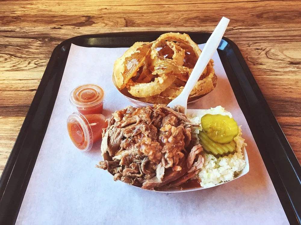 BBQ tray with white slaw, pickles, pulled pork, and onion rings on a black cafeteria tray