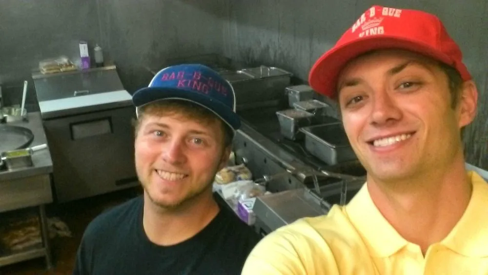 Jordan and Jared Smith smiling for a selfie in the BBQ King kitchen wearing Bar-B-Que King caps