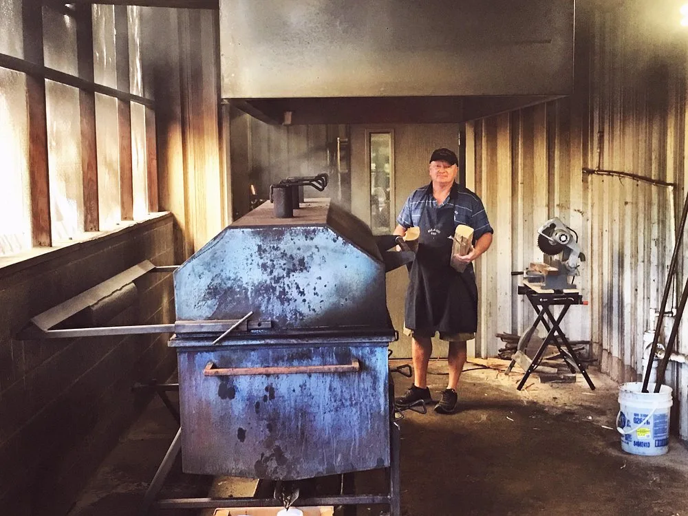 Keith Smith standing beside the pit smoker with a chop saw and stacked hickory wood behind him
