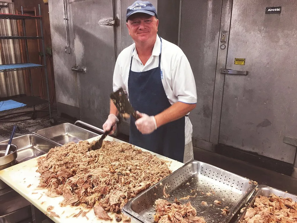 BBQ King team member in a Bar-B-Q King cap hand chopping a large pile of pulled pork by the walk-in coolers