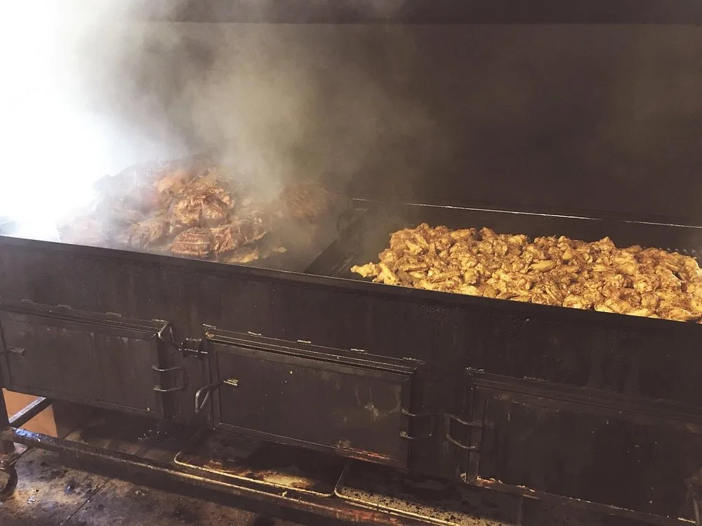 Pork shoulders and seasoned chicken smoking side by side on the open pit, wrapped in hickory smoke