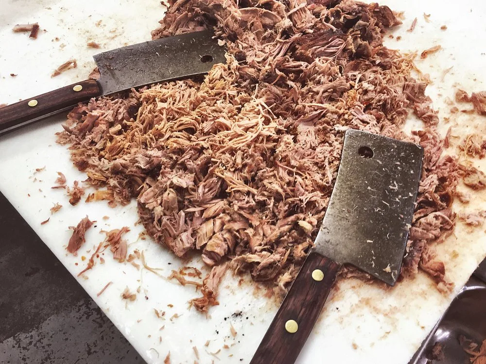 Two well-worn meat cleavers resting on a cutting board next to a pile of freshly pulled pork