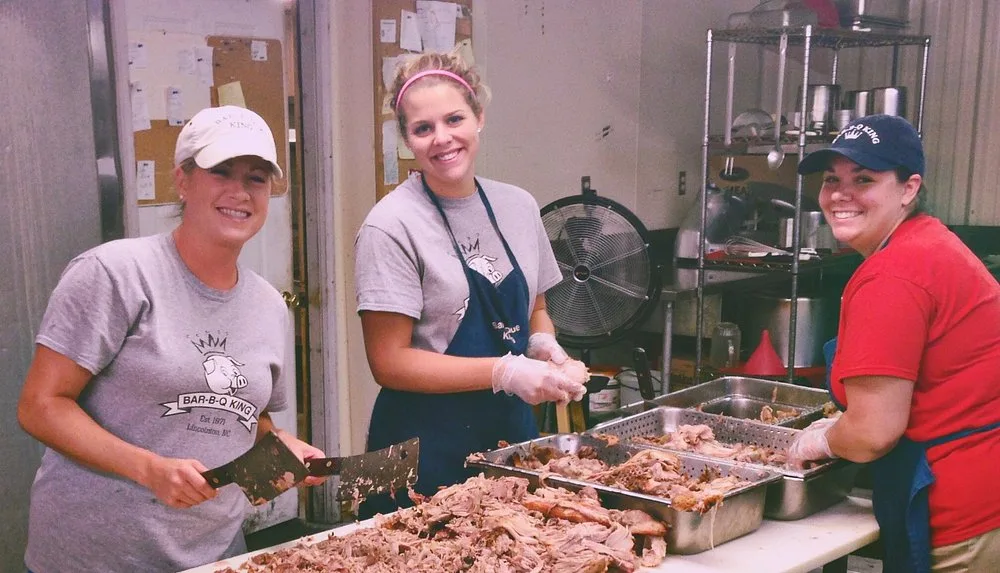 Three BBQ King employees in matching Bar-B-Q King shirts chopping pork together in the smokehouse kitchen