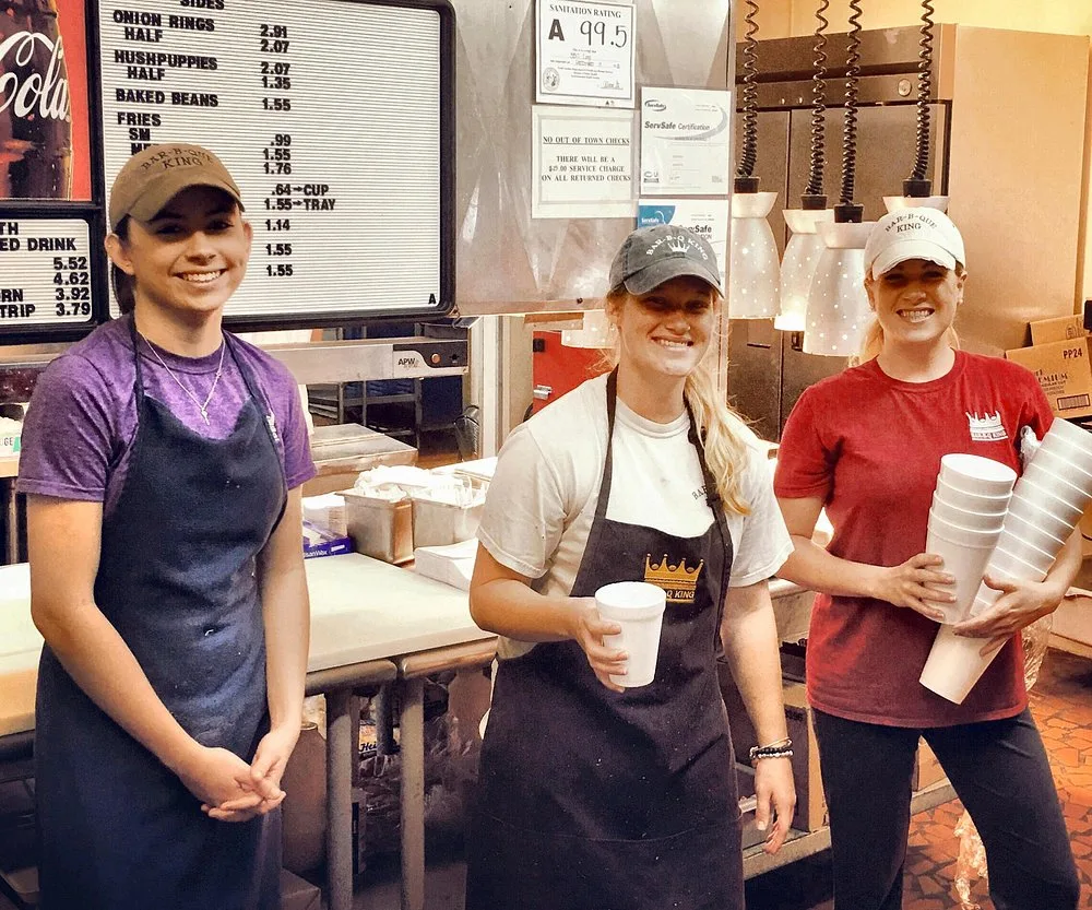 Three BBQ King counter staff in Bar-B-Q King caps smiling in front of the menu board