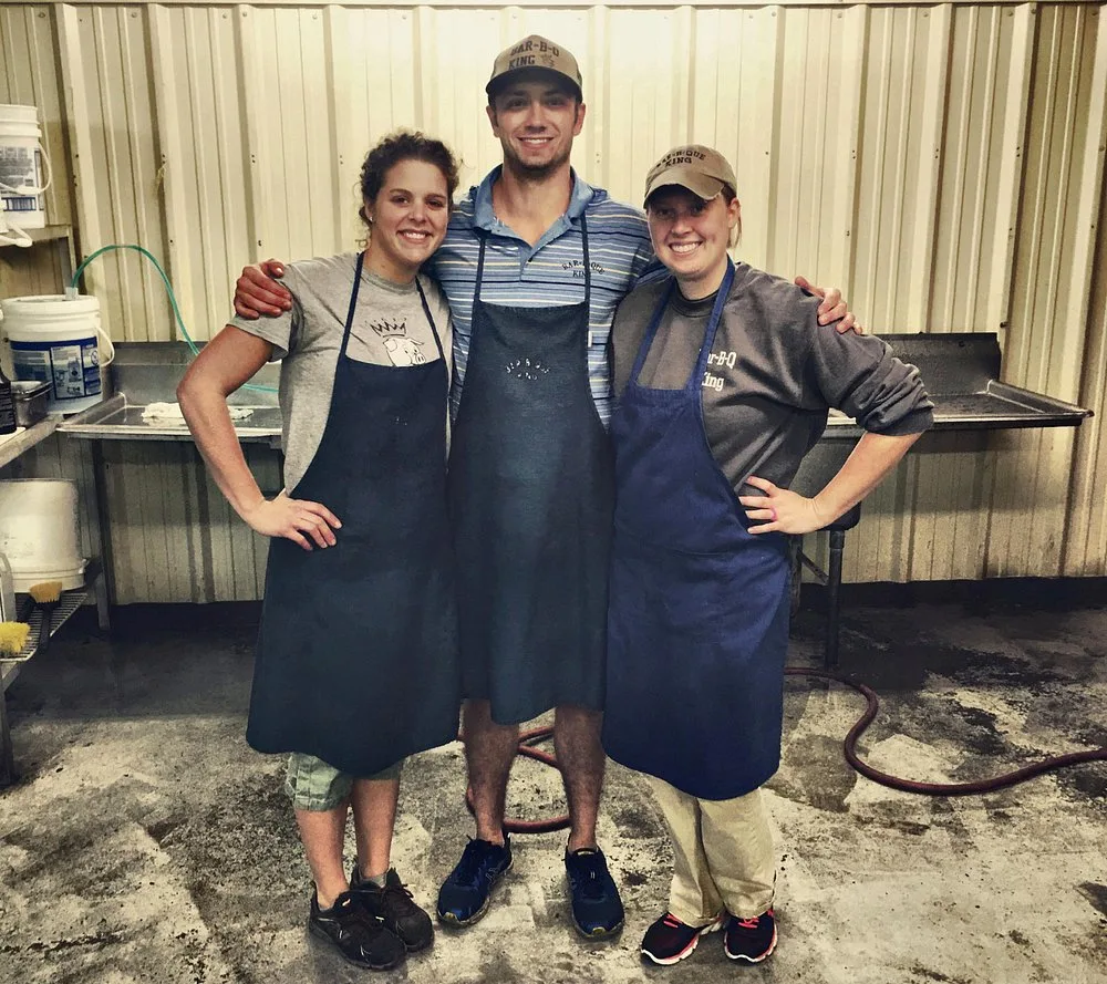 Three BBQ King employees in navy aprons posing together in the prep kitchen