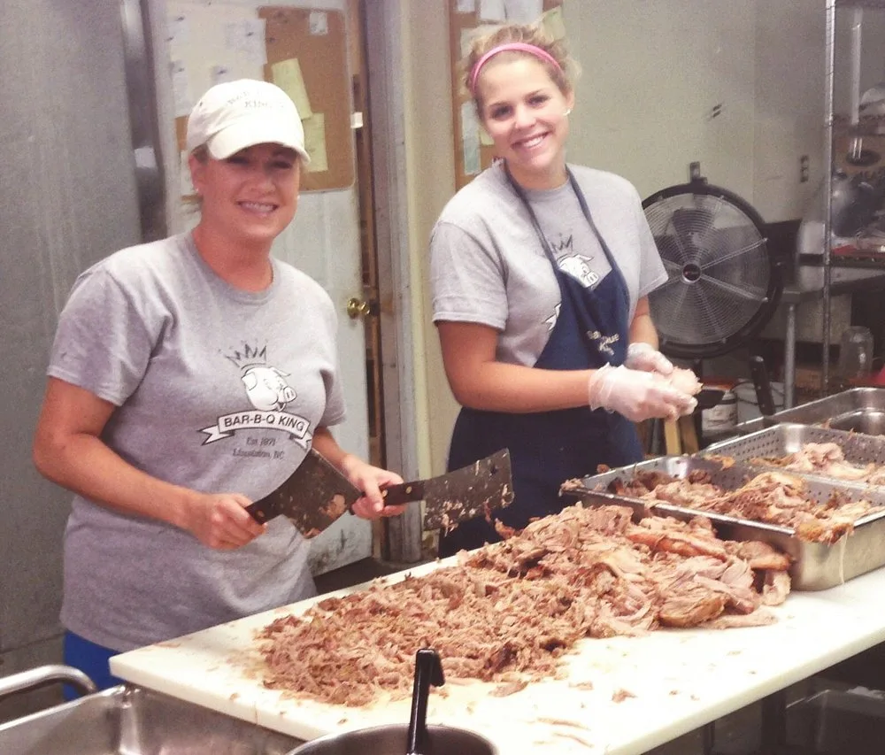 Two smiling BBQ King employees holding cleavers over a large pile of pulled pork