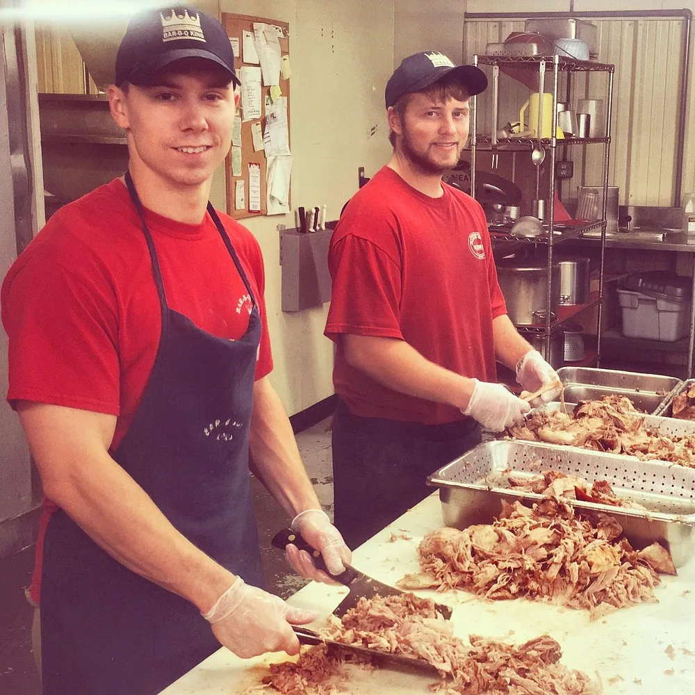 Two BBQ King employees in red shirts pulling pork into steel prep pans
